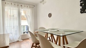 Un comedor con mesa de vidrio, sillas blancas, ventilador de techo y vista a un balcón con plantas.