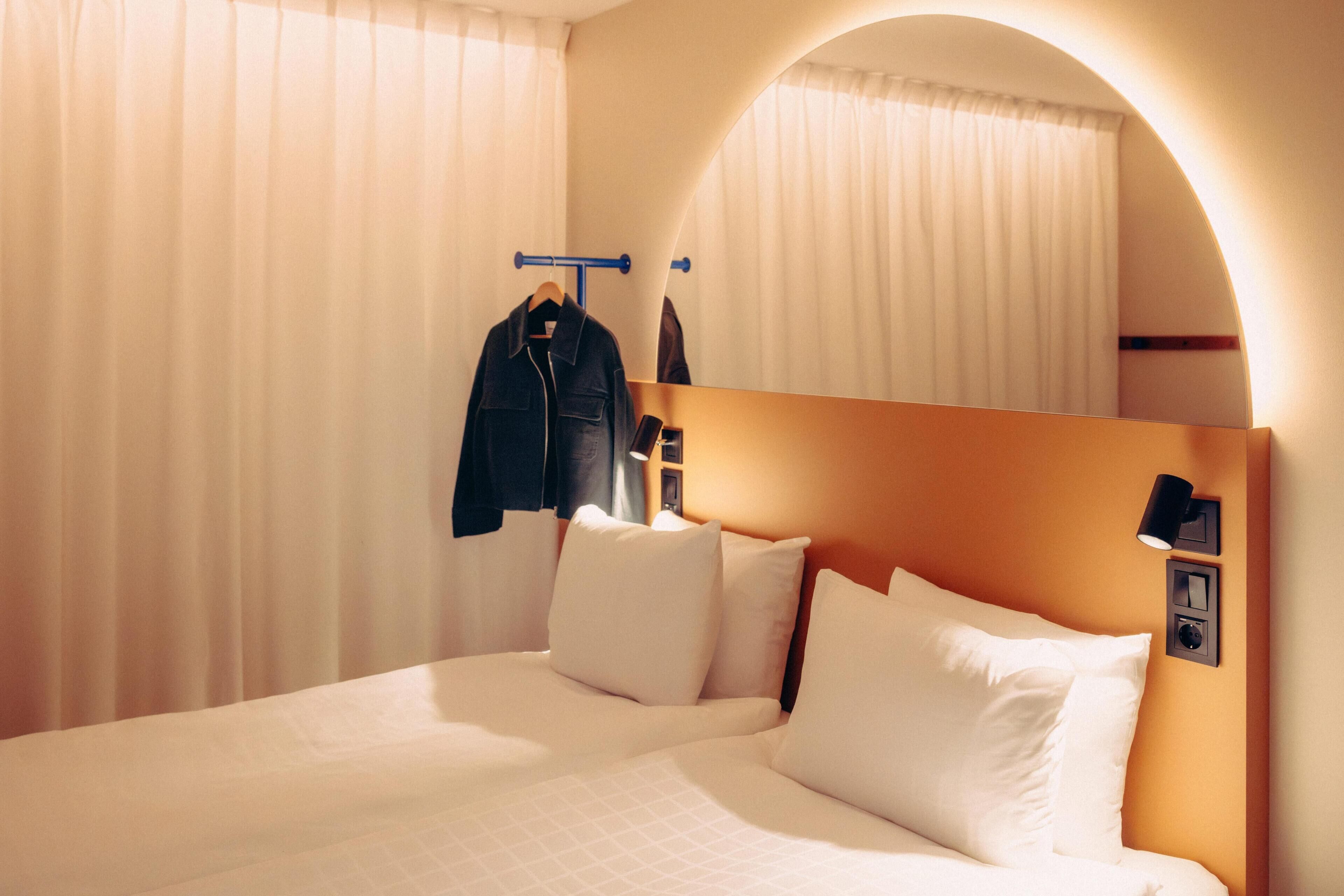 Habitación de hotel con cama, cortinas blancas, perchero y lámpara fijada a la pared.