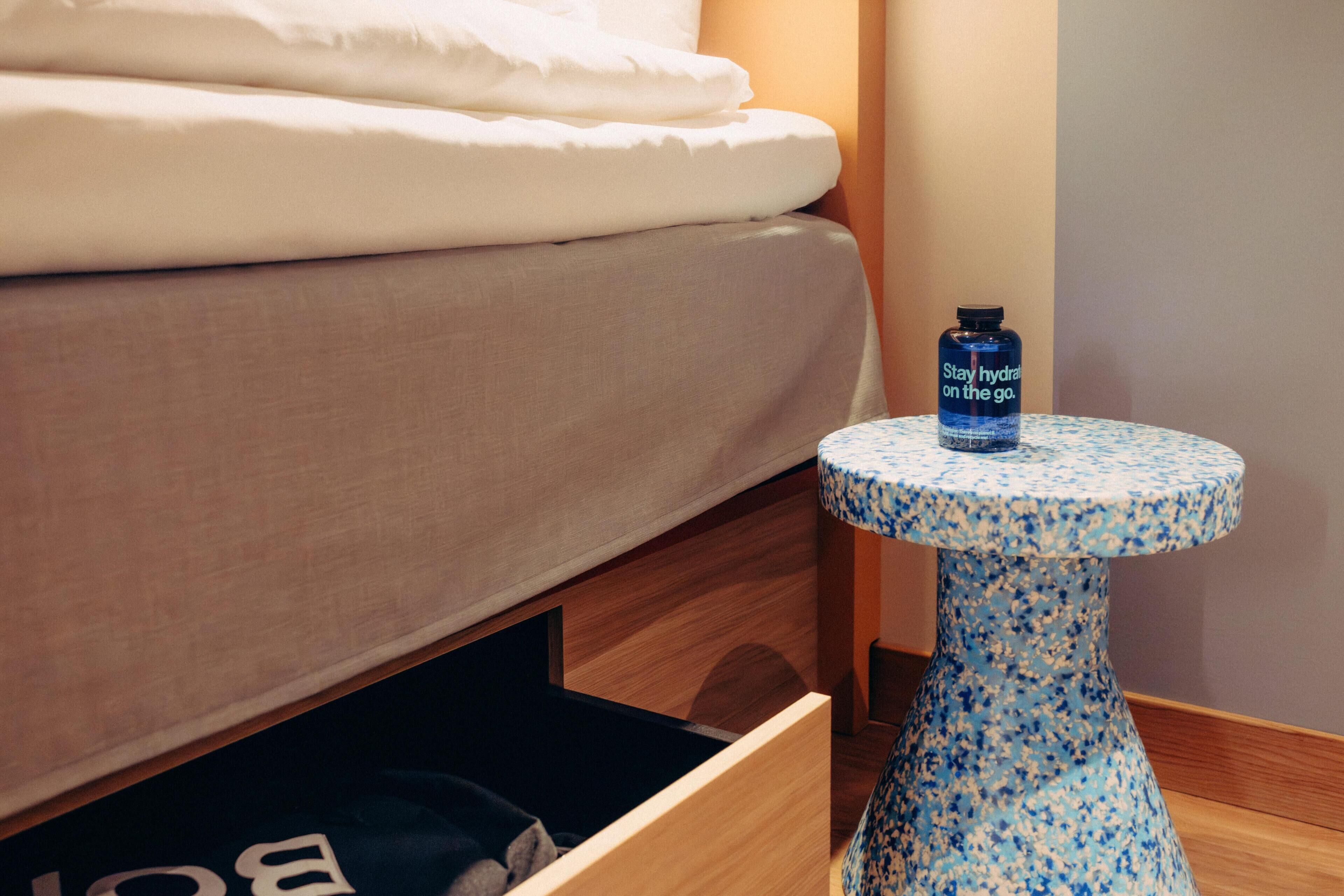 A bed with a white comforter, a small blue table with a bottle, and a drawer with a black item.