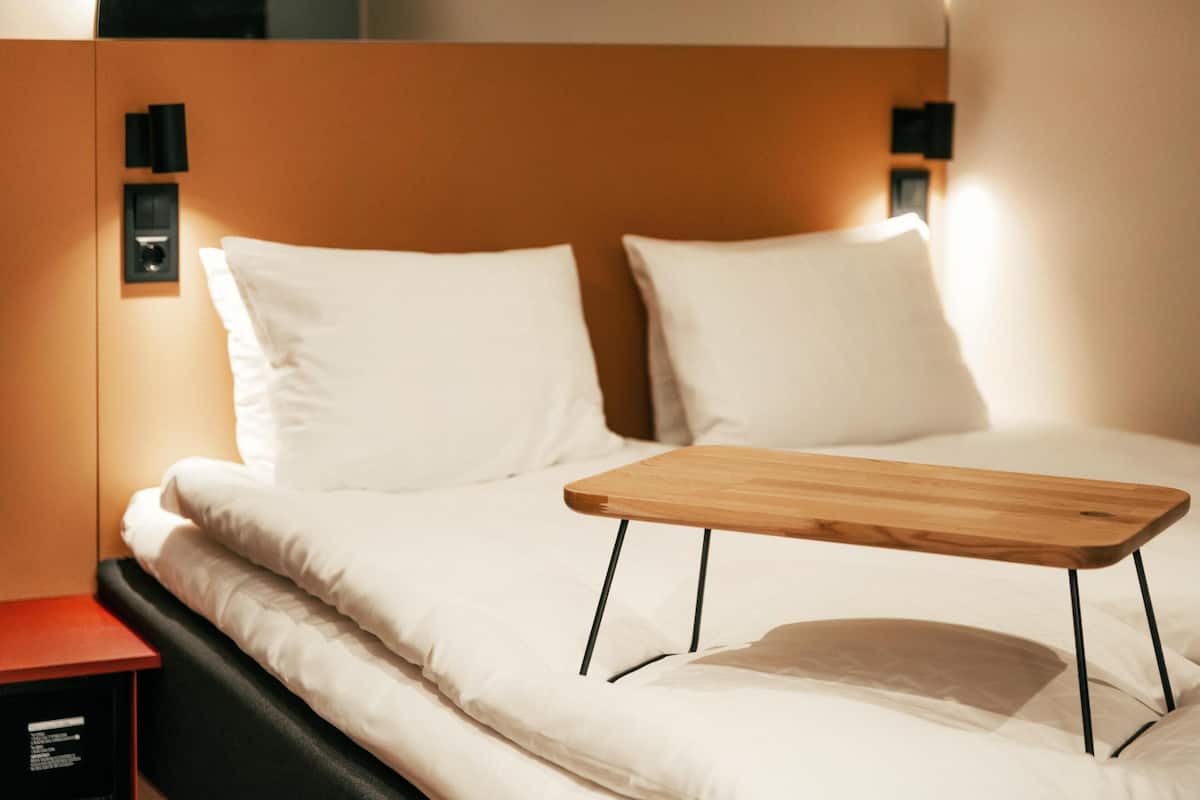 Una cama bien hecha con sábanas blancas, una mesita de noche de madera y lámparas negras fijadas a la pared.