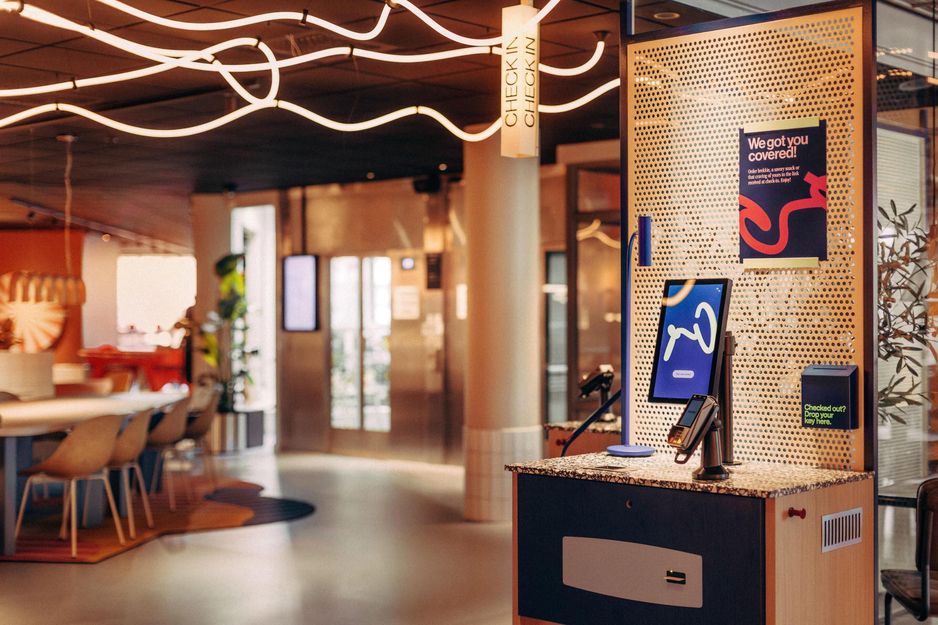 Un mostrador de recepción moderno con un quiosco digital, un cartel que indica el registro y una estación de devolución de llaves fijada a la pared.