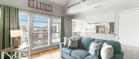 A modern living room with a blue sofa, a small side table with a lamp, and a large window with blinds.