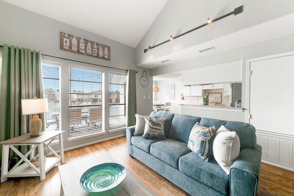 A modern living room with a blue sofa, a small side table with a lamp, and a large window with blinds. - Waterfront Condo | Spacious Deck & Intracoastal Waterway Access (Corpus Christi)