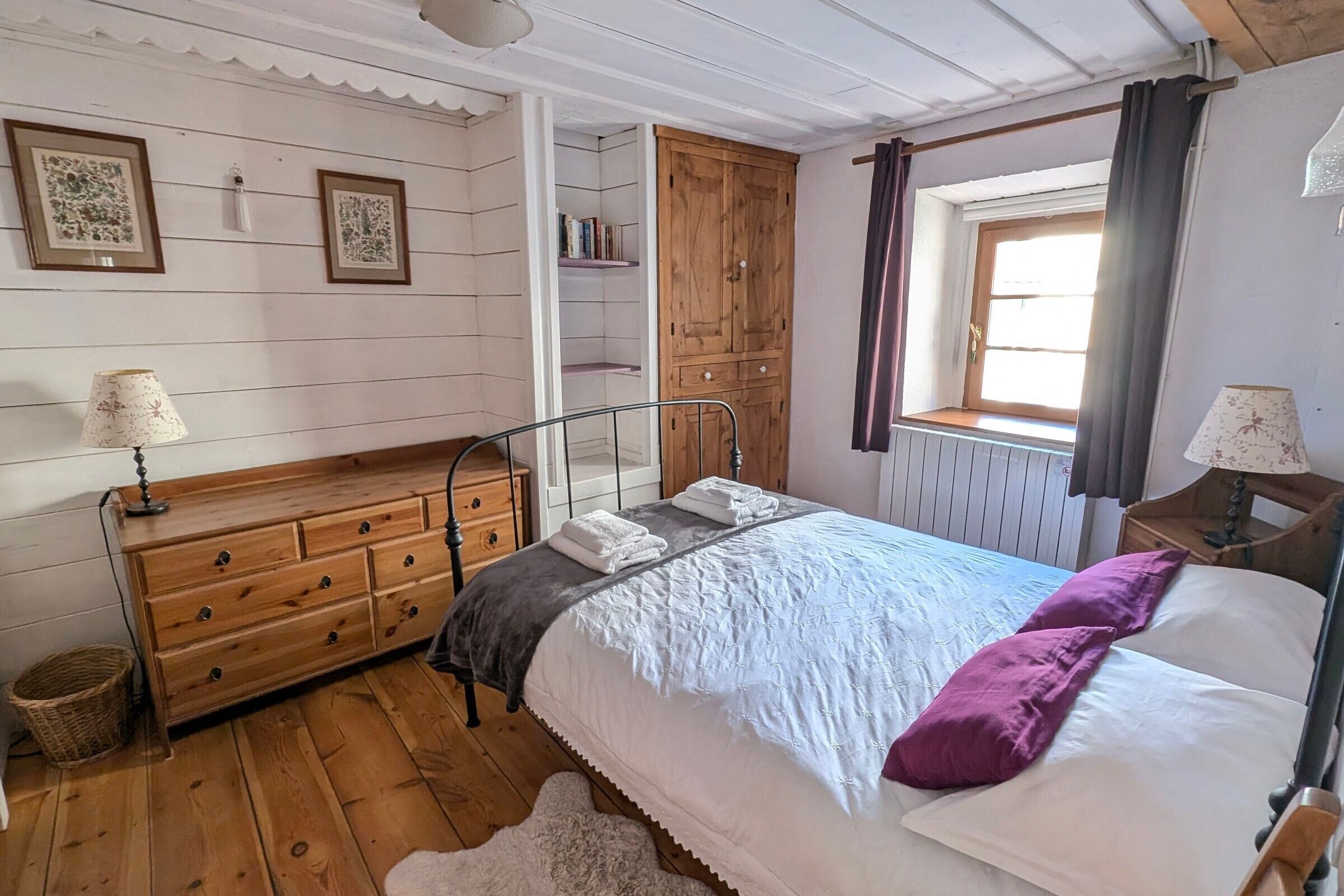 A bedroom with a wooden dresser, a bed with white and purple bedding, a window with curtains, and a wooden wardrobe.
