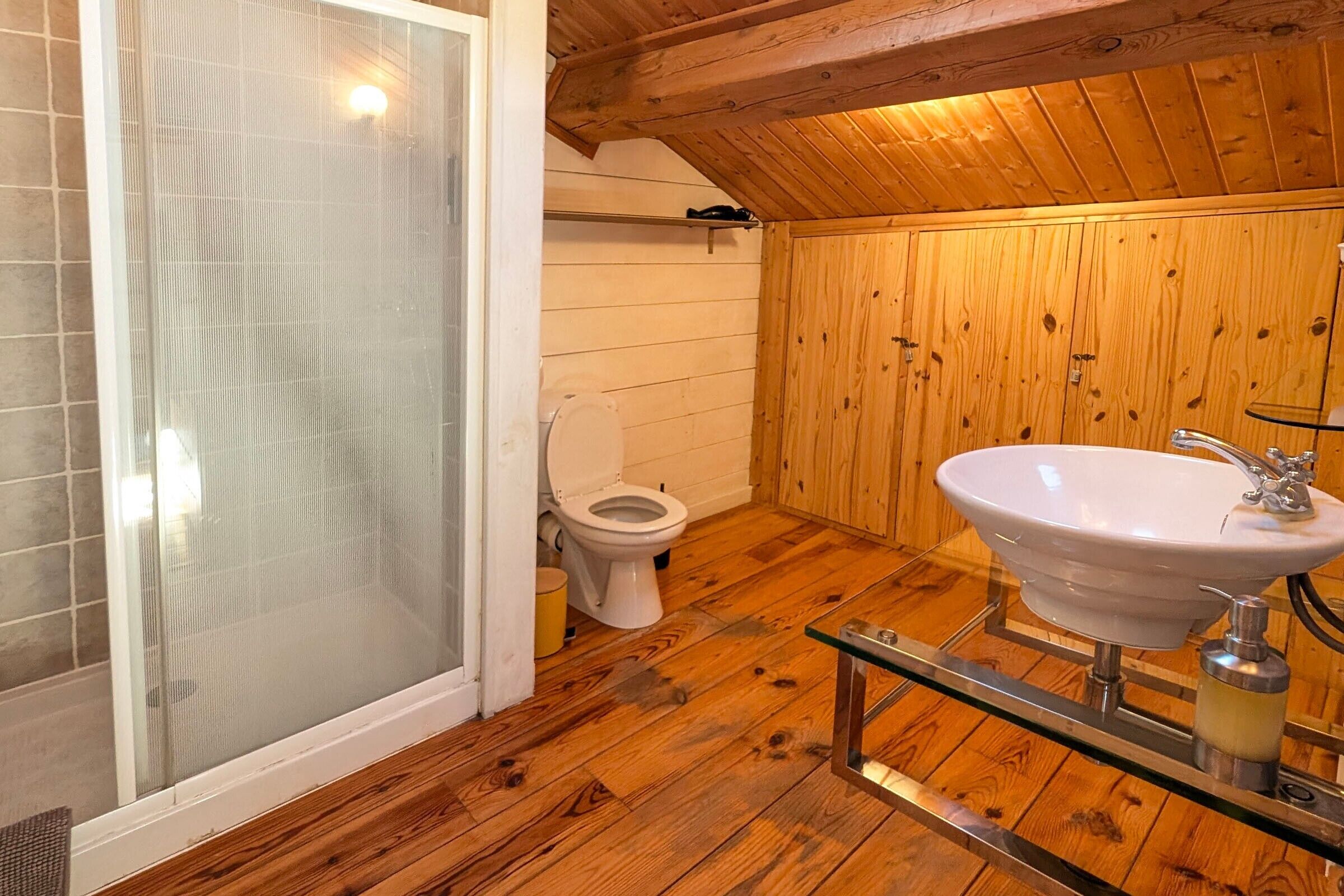 Un baño con paneles de madera, cabina de ducha de vidrio, inodoro y lavamanos blanco con estante de vidrio.
