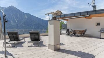 A terrace with two lounge chairs, a table, and a grill, overlooking a mountain and a body of water.