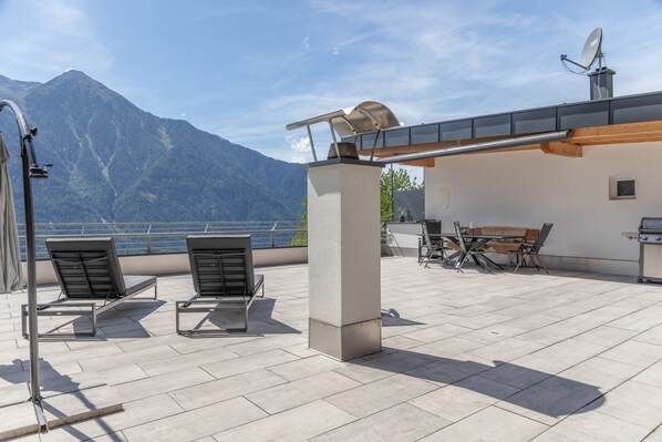 A terrace with two lounge chairs, a table, and a grill, overlooking a mountain and a body of water.