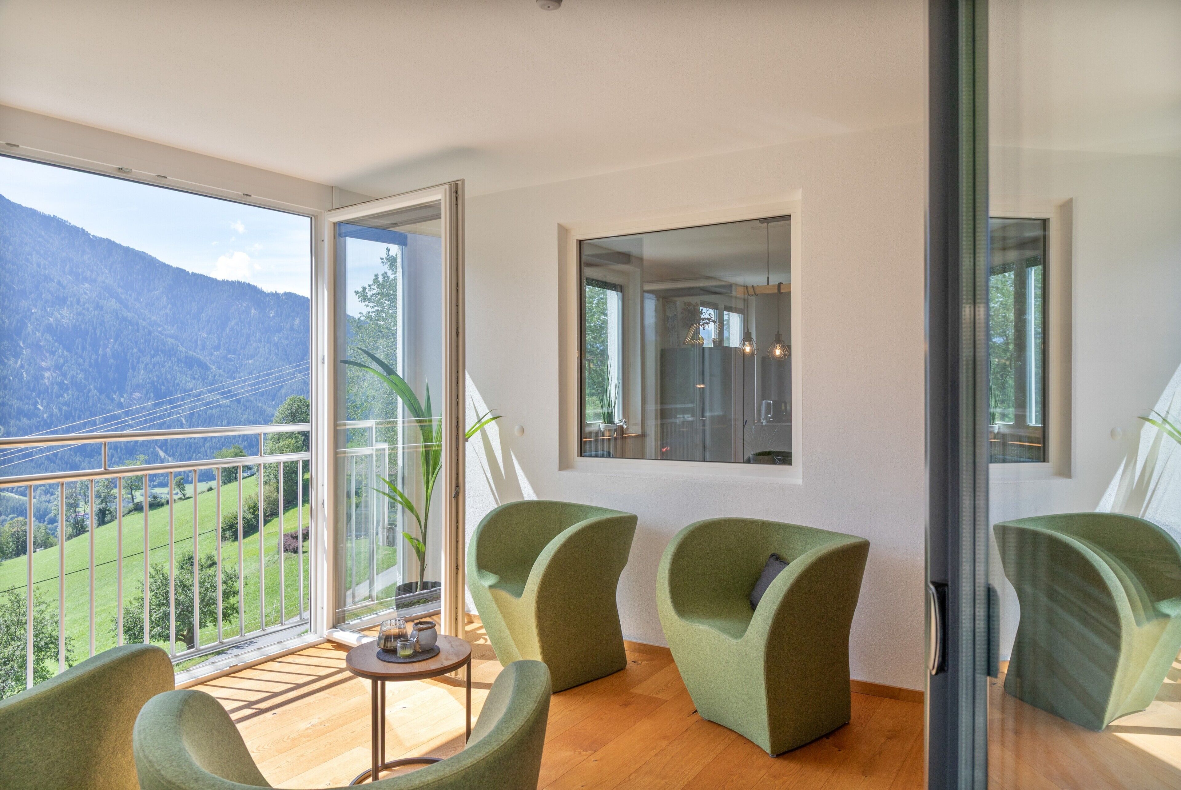 A room with a balcony, three green chairs, a small table, and a view of a mountainous landscape.