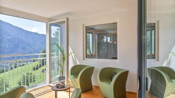 A room with a balcony, three green chairs, a small table, and a view of a mountainous landscape.