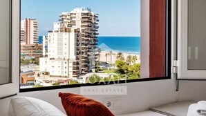 Quarto de hotel com uma grande janela que oferece vista para uma praia e edifícios altos.
