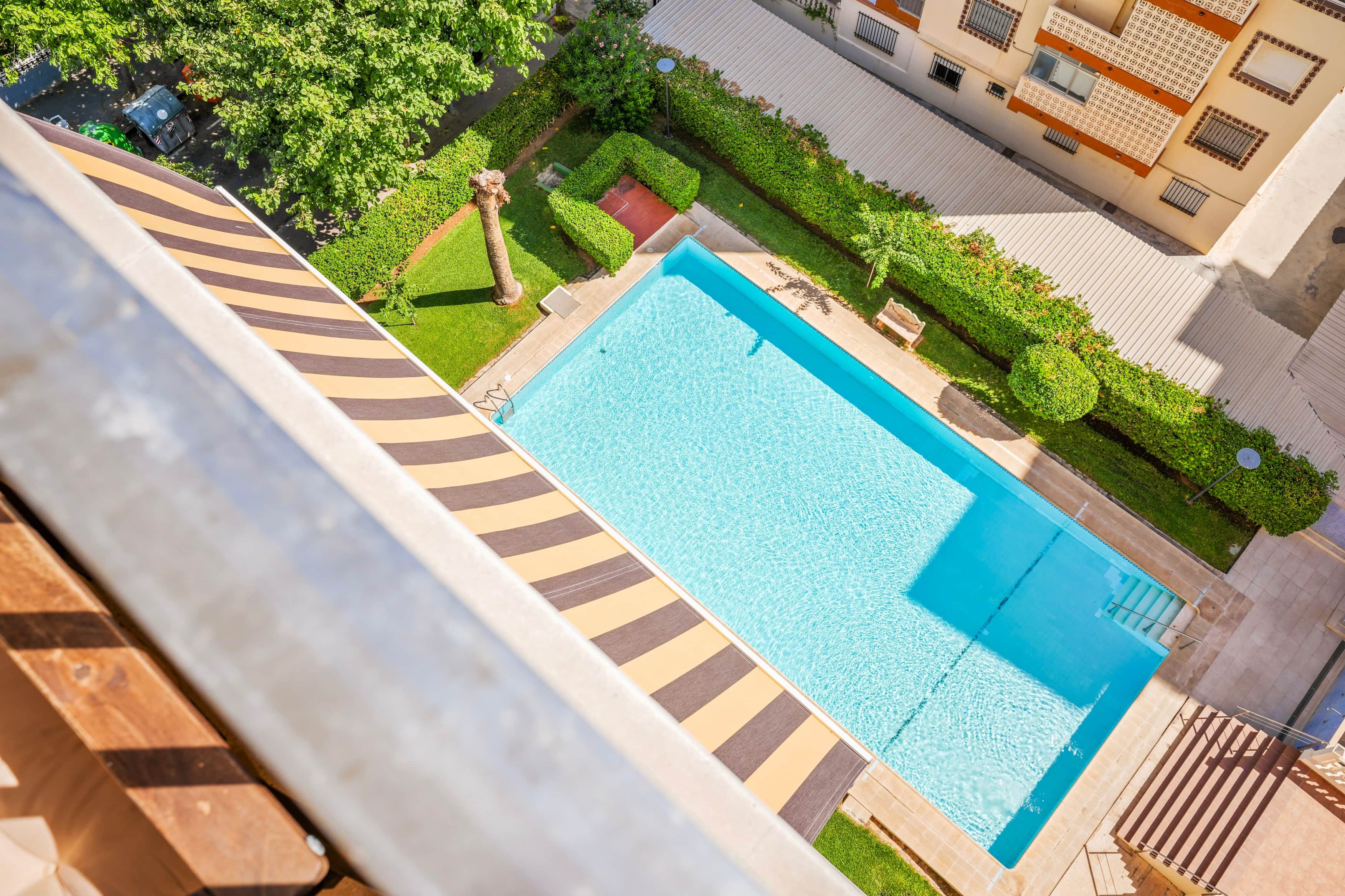 Una piscina rettangolare con superficie d'acqua cristallina e blu, circondata da un giardino curato e piante varie, con un vialetto lastricato.