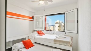 Quarto de hotel moderno com duas camas, ventoinha no teto e uma janela grande com vista para a cidade.