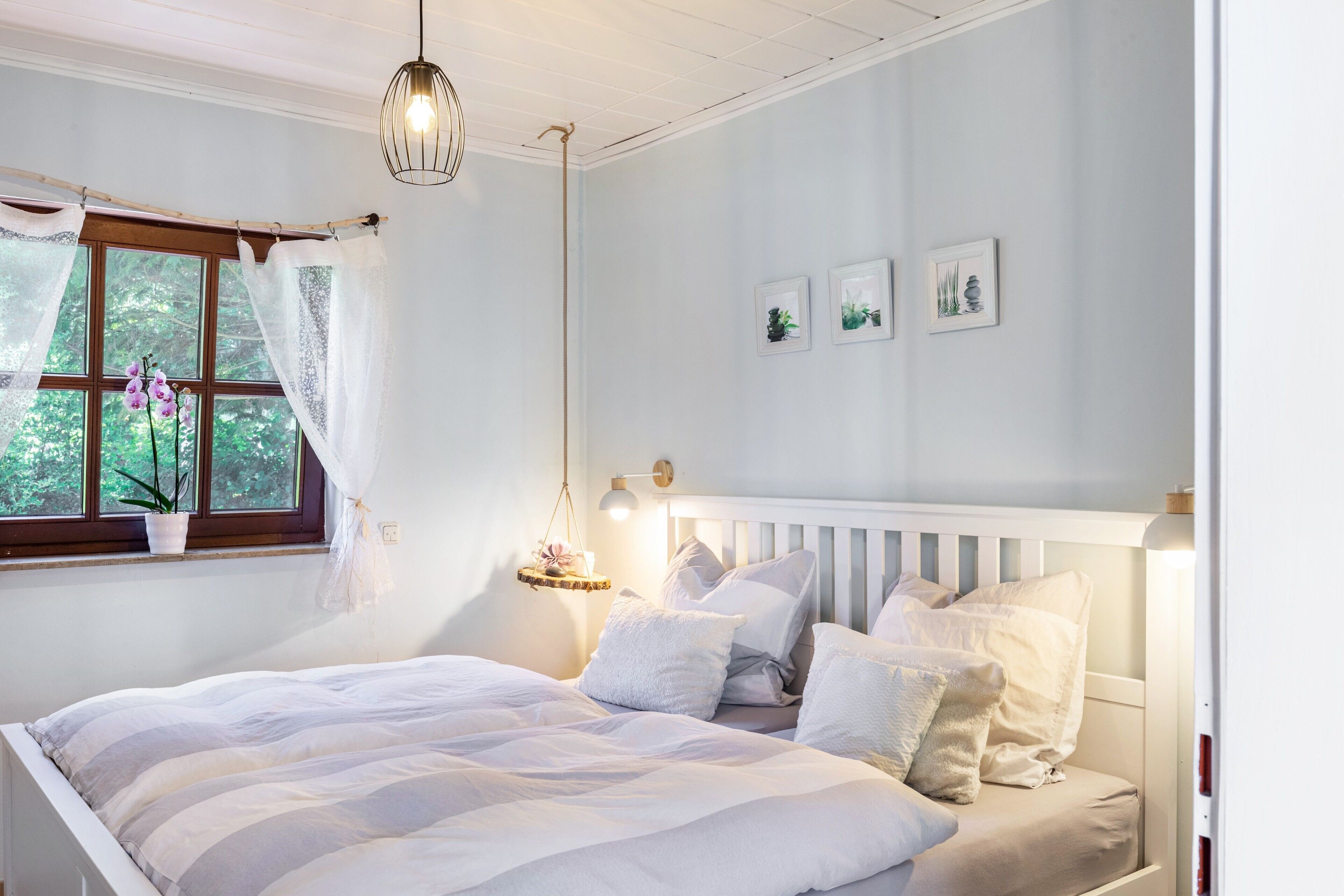 Ein Schlafzimmer mit einem Bett, einem Fenster mit Vorhängen, einer Hängelampe und an der Wand befestigten Bildern.
