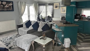 A compact living space with a sofa, a small table, a kitchen area, and a dining table. - Caravan - Sleeps 6 - Short Walk to Rhyl Beach (Rhyl)