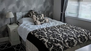 A bedroom with a bed, a nightstand, a lamp, a window with blinds, and a patterned wall. - Caravan - Sleeps 6 - Short Walk to Rhyl Beach (Rhyl)