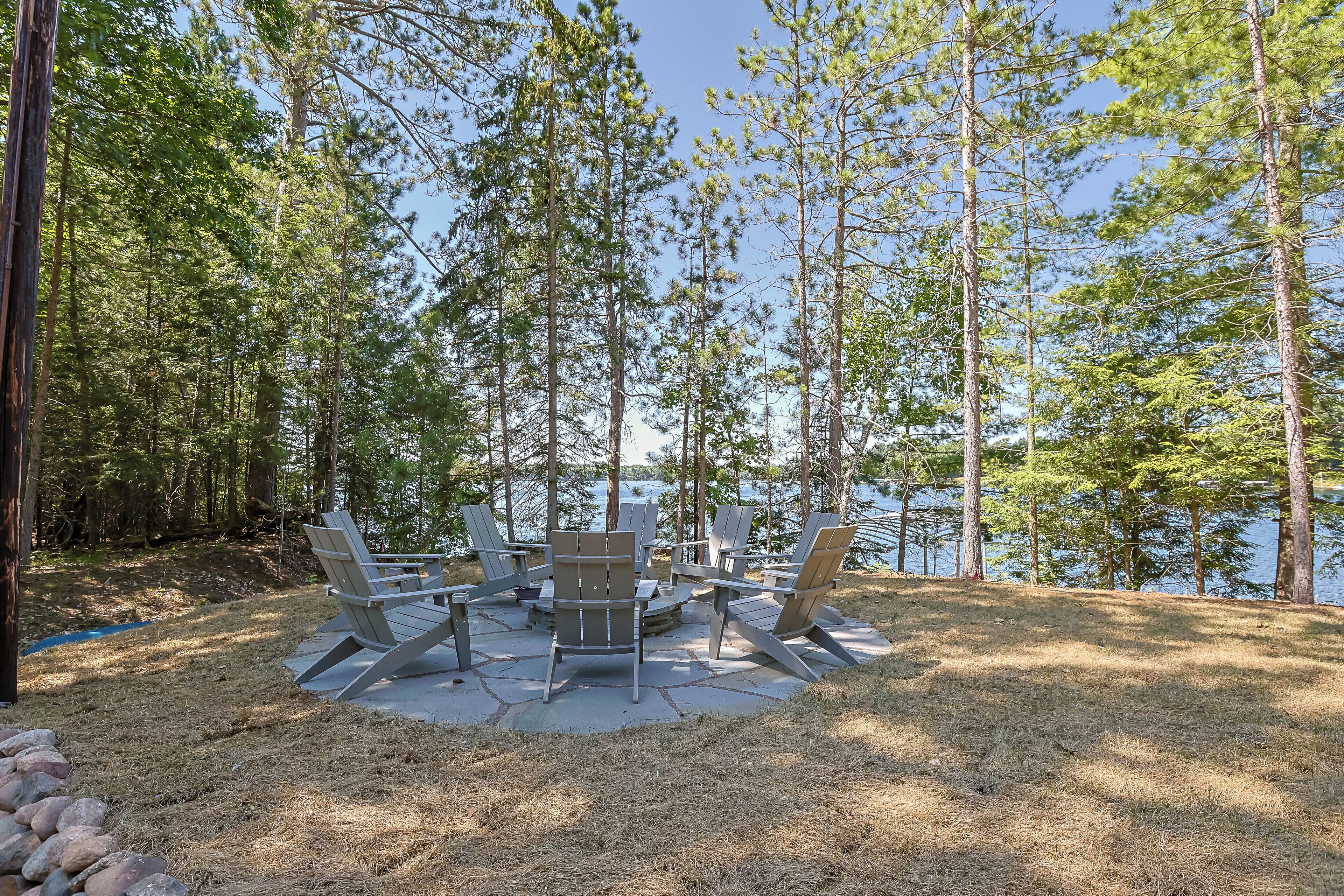 Un espacio de estar circular al aire libre con sillas Adirondack alrededor de una mesa central, rodeado por un paisaje natural con árboles altos y un cuerpo de agua al fondo.