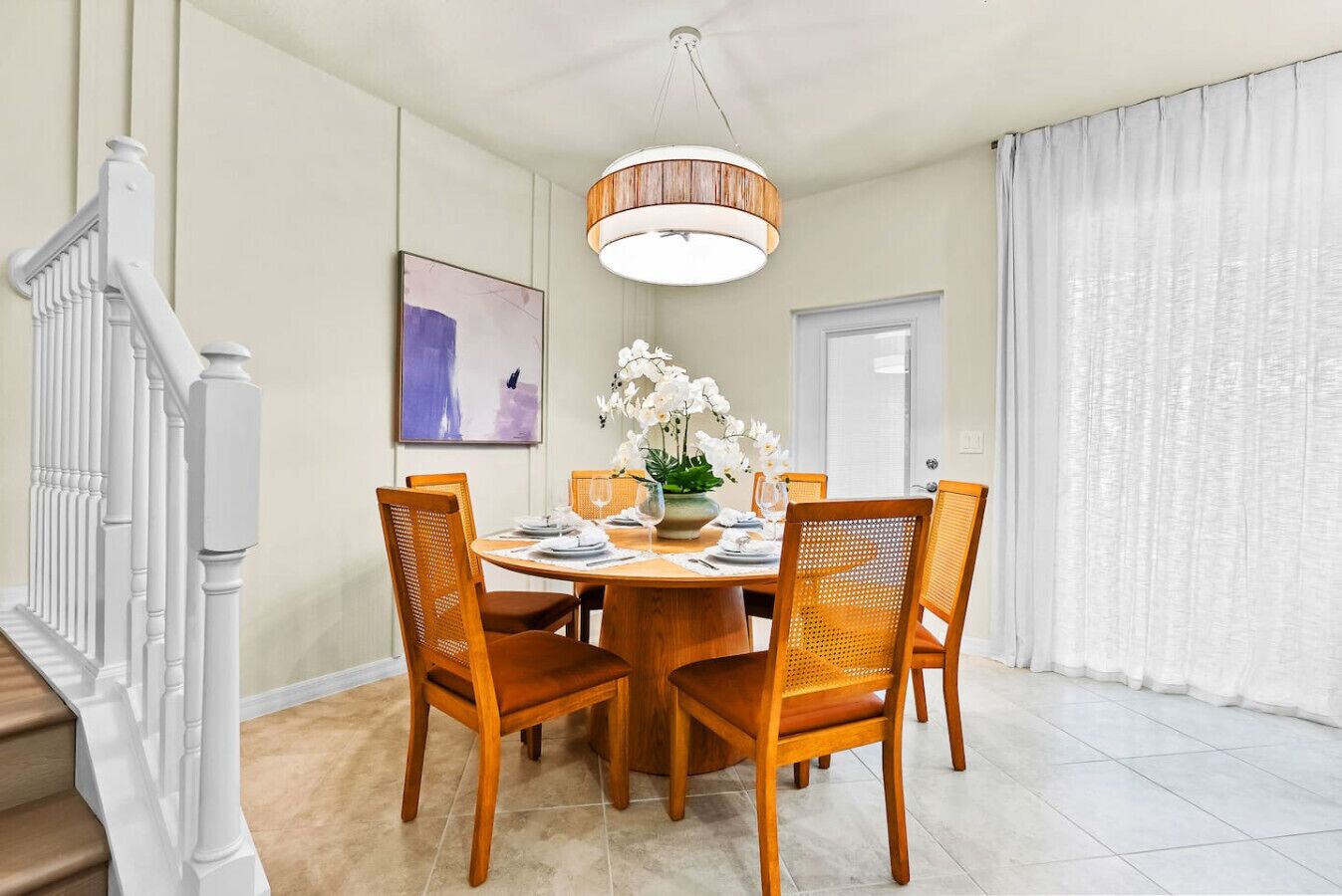 A dining area with a round table, four wooden chairs, a vase of white flowers, and a modern pendant light.