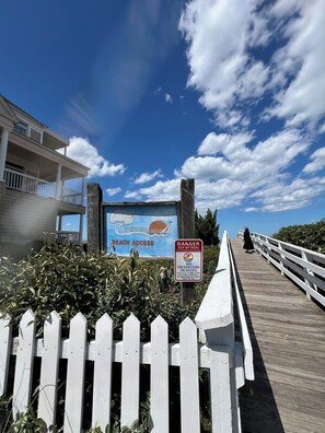 Ein hölzerner Steg mit einem Warnschild, einem Schild für den Zugang zum Strand und einem Gebäude mit einem Balkon.