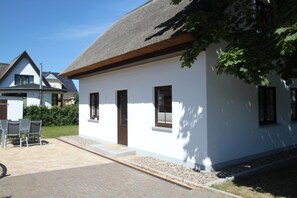 Un bâtiment blanc avec un toit de chaume, une porte et des fenêtres. On y trouve une zone pavée avec des chaises et une table.