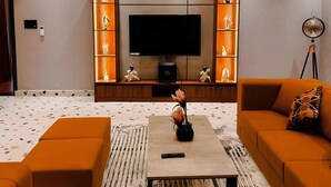 Un salon moderne doté d’un grand téléviseur à écran plat, de canapés orange, d’une table basse agrémentée d’un vase et d’un lustre.