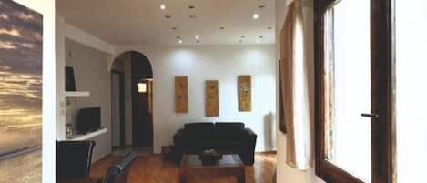 A spacious hallway with wooden flooring, a black sofa, a coffee table, and a dining area with chairs. There are ceiling lights and a window on the right.