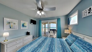Ein Schlafzimmer mit Bett, Deckenventilator, Kunstwerken, Fernseher und Blick auf den Strand.