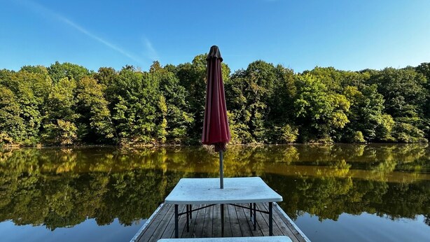 Un molo di legno che si protende su un lago tranquillo, con un ombrellone rosso e una panchina.