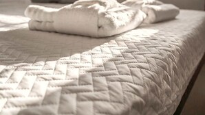 Ein ordentlich bezogenes Bett mit einer gesteppten Tagesdecke, ein Fenster mit Vorhängen und eine Nachttischlampe.