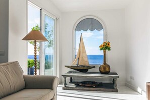 Ein Wohnzimmer mit einer Couch, einer Lampe, einem Tisch mit einem Modell eines Segelboots, einer Vase mit Blumen und Meerblick durch ein Fenster.