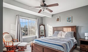 A bedroom with a bed, a chair, a ceiling fan, a window, and artwork on the wall.