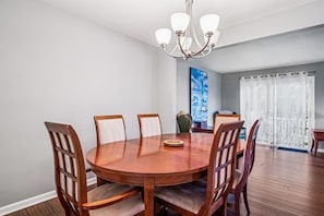 A dining area with a round wooden table, six chairs, a chandelier, and a large window with curtains.