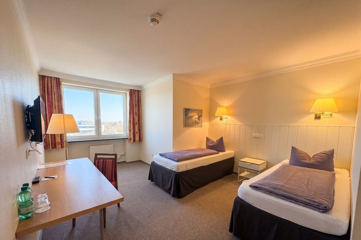 A hotel room with two beds, a desk, a chair, a TV, and a window with curtains.
