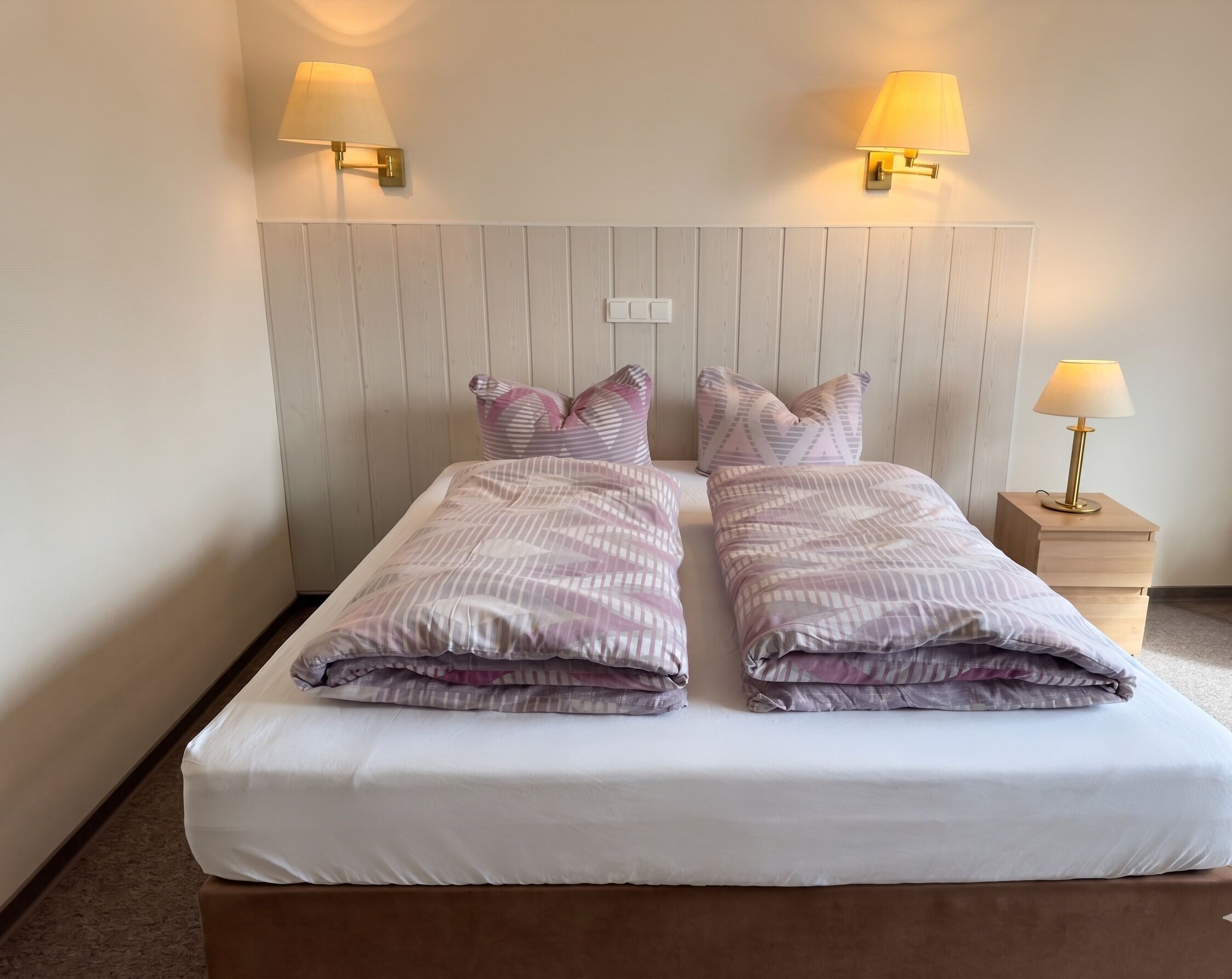 Een tweepersoonsbed met een witte matras en twee kussens, een houten hoofdbord en twee wandlampen.