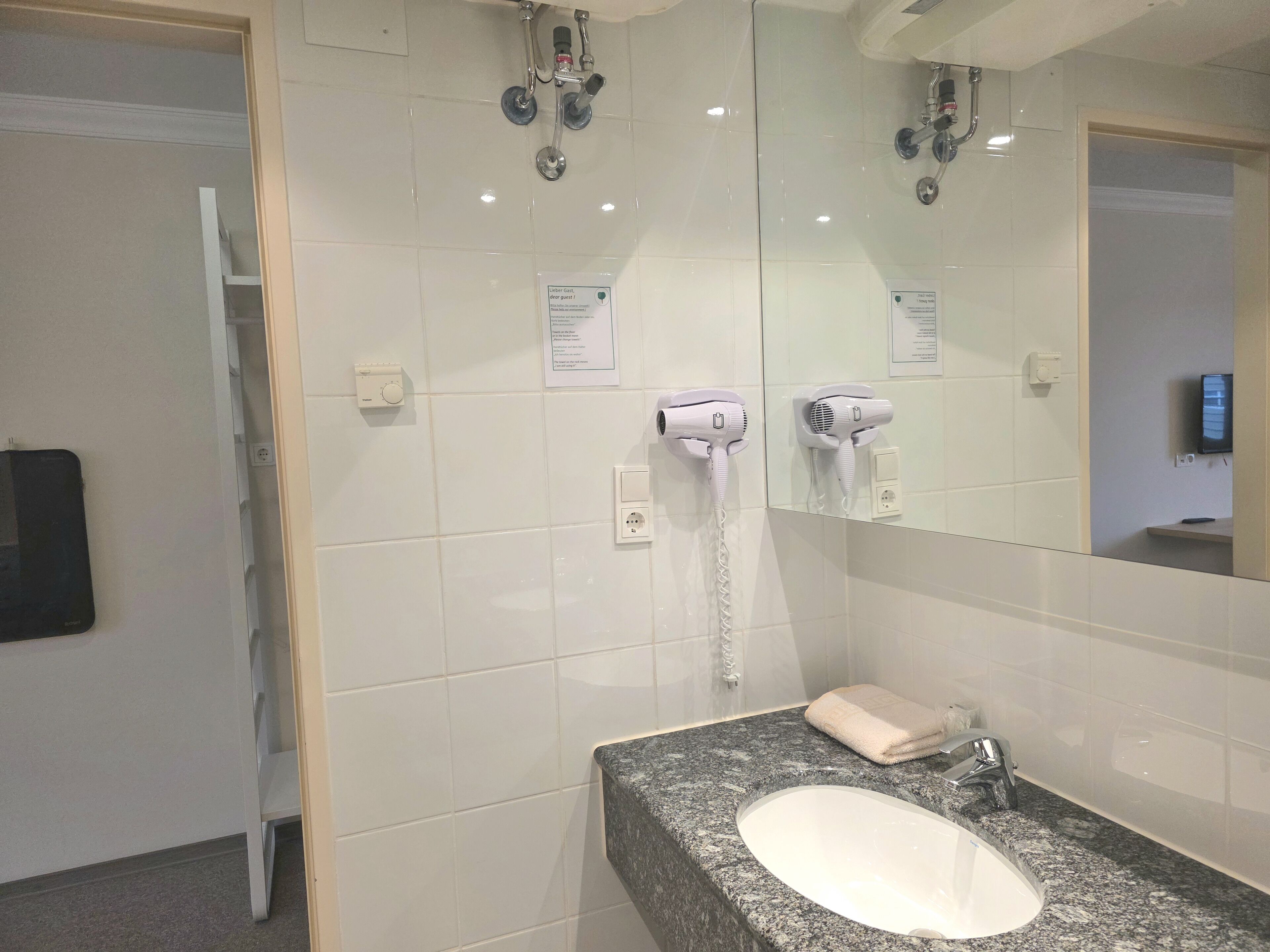 A hotel bathroom with a granite countertop sink, a towel, a mirror, and two hair dryers mounted on the wall.