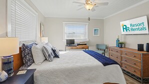 Een slaapkamer met een bed, een bureau, een dressoir, een televisie en een plafondventilator.