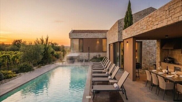 A poolside area with lounge chairs and a dining setup, overlooking a landscaped garden and a building with a stone facade. - Deluxe Villa Brtonigla with heated pool (Brtonigla)
