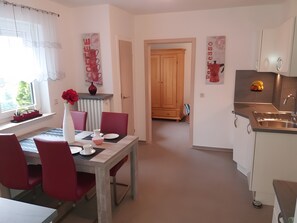 A dining area with a table set for two, red chairs, a vase with flowers, and a kitchen with a sink and cabinets.