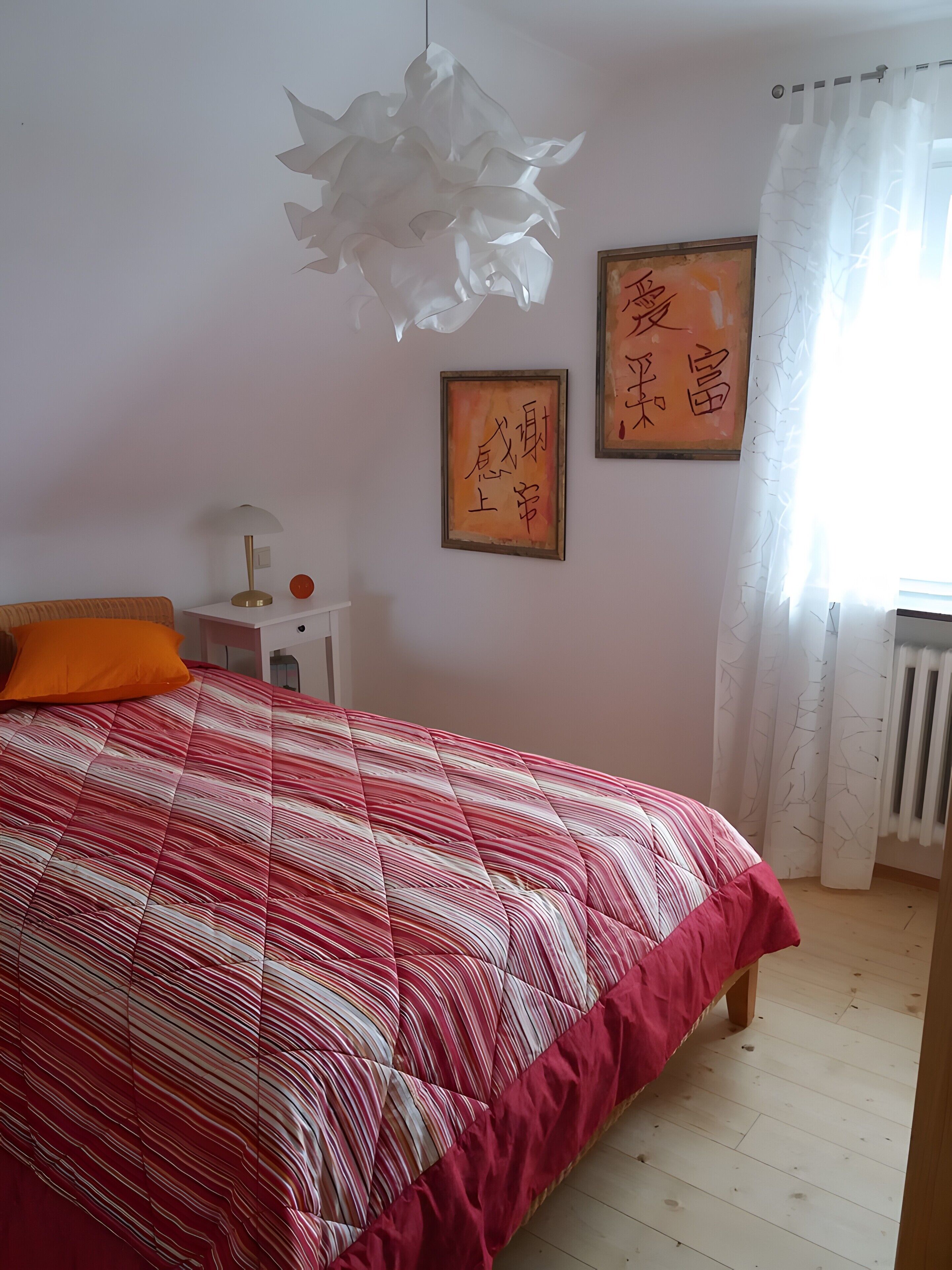 A bedroom with a bed, a nightstand, a lamp, two framed calligraphy pieces, and a window with curtains.