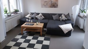 Un salon moderne avec un sol à carreaux, un canapé gris agrémenté de coussins à motifs, une table basse en bois ornée d’objets décoratifs et un tableau accroché au mur.