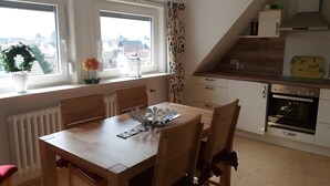 Un coin repas avec une table et des chaises en bois, une vue par la fenêtre, une cuisine équipée d’une cuisinière et de placards, et un radiateur.