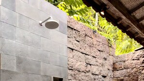 Um chuveiro moderno ao ar livre, com parede de pedra ao fundo, prateleira pequena com produtos de higiene pessoal e um caminho revestido de pedrinhas.