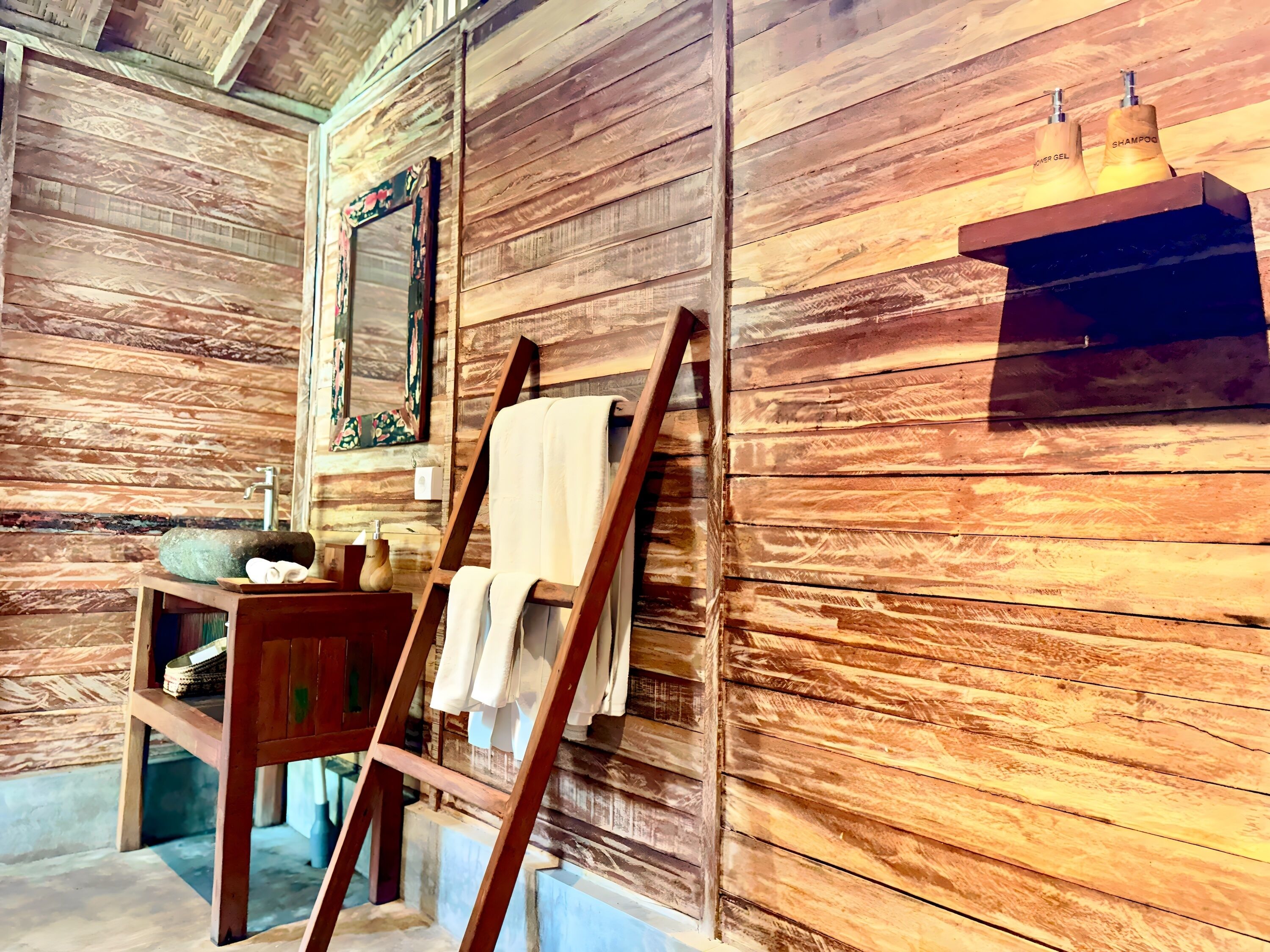 A rustic bathroom with wooden walls, a wooden bench, a mirror, a shelf with toiletries, and a towel rack.