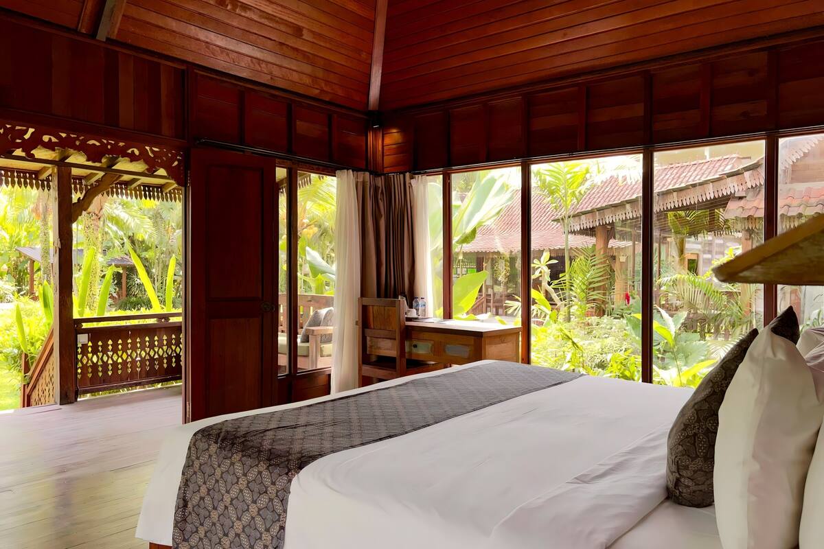 Una habitación de madera con una cama, ventanas amplias y vistas a un jardín tropical.