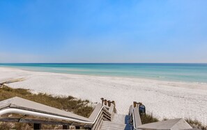 Ein Strand mit einer Holztreppe, die zum Sand führt, unter blauem Himmel und ruhigem Meer.
