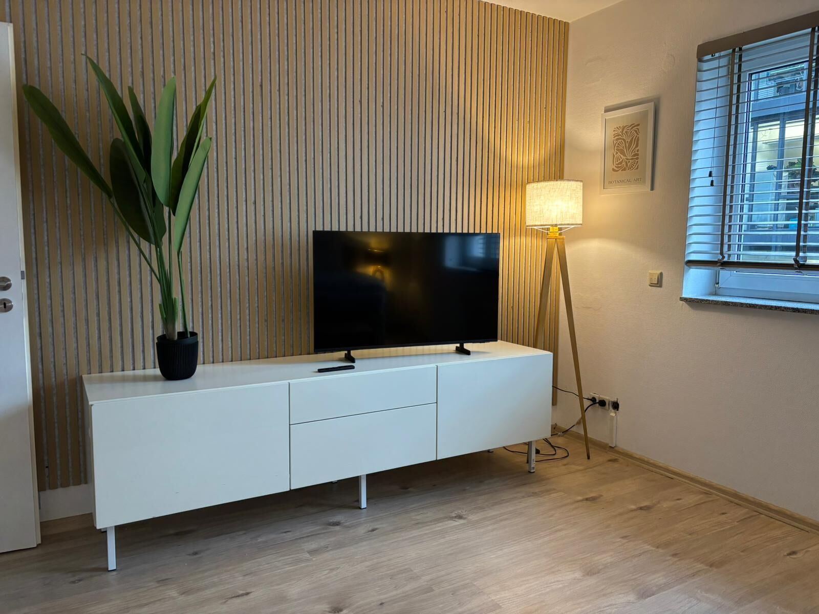 Una sala de estar moderna con un televisor de pantalla plana sobre un mueble blanco, una planta en maceta, una lámpara de pie y una ventana con persianas.