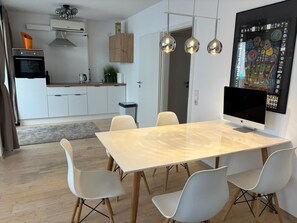 A modern kitchen with white cabinets, a built-in oven, and a coffee machine. There is a dining area with a large table and white chairs. - Modern Apartment | Karlsruhe City Center (Karlsruhe)