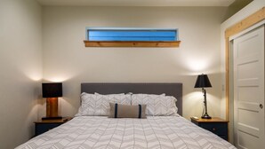 Una cama bien hecha con una colcha estampada, dos lámparas de noche, una ventana con marco de madera y una puerta a la derecha.