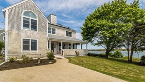 A two-story house with a covered porch, a landscaped yard, and a view of a lake. - Quintessential Cape Cod home on Great Pond RPLO026 (Teaticket)