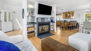 A modern living room with a fireplace, white sofas, wicker ottomans, and a dining area with wooden chairs. - Quintessential Cape Cod home on Great Pond RPLO026 (Teaticket)