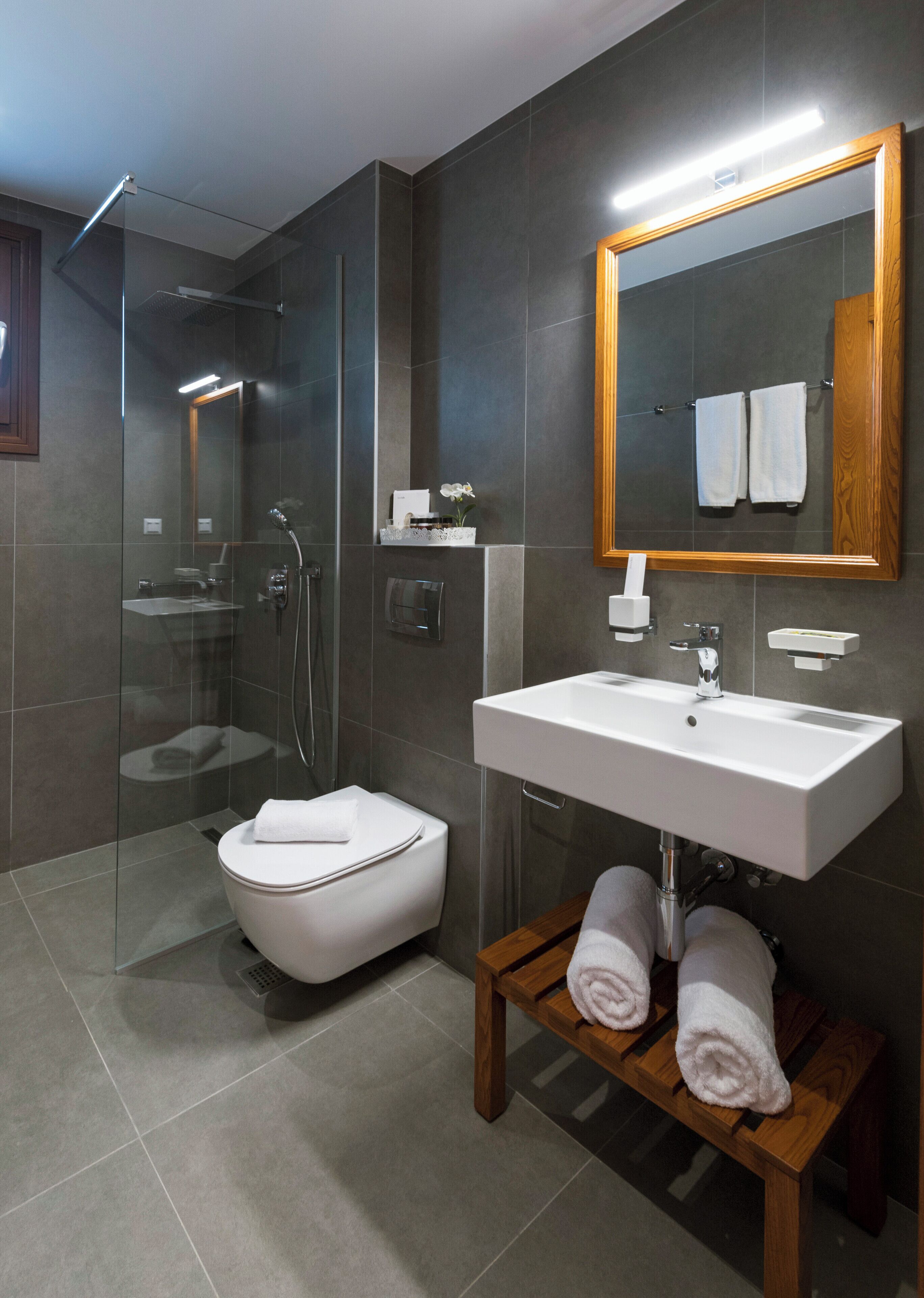 Un bagno moderno con doccia vetrata, un water bianco, un mobile bagno in legno con lavandino e uno specchio con cornice in legno.